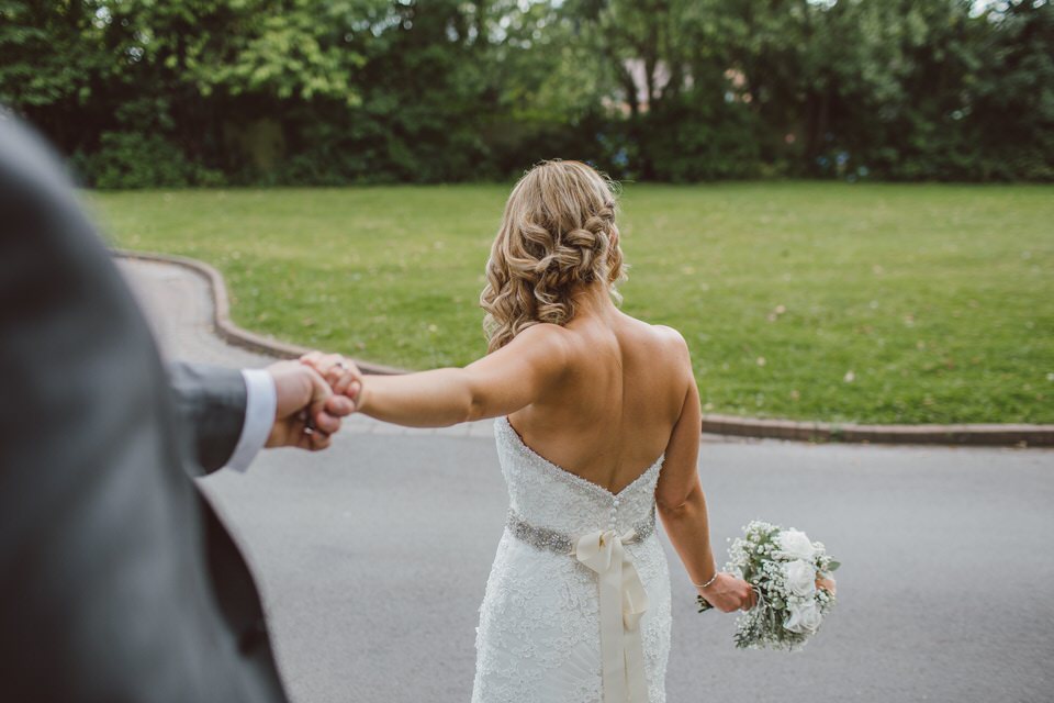 A photo of a new bride holding her husband's hand in a column shaped wedding dress